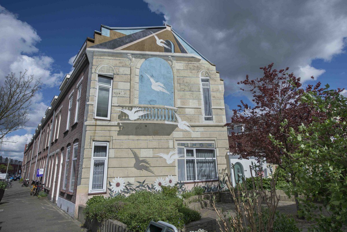 Muurschildering toont een gevel met ramen en een balkon, waarvoor enkele duiven in vlucht zijn afgebeeld.
